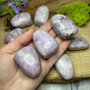 May include: A selection of tumbled lilac-coloured stones with white marbling. The stones are smooth and oval-shaped, with varying shades of purple. The stones are displayed on a wooden surface, with green moss and purple flowers in the background.