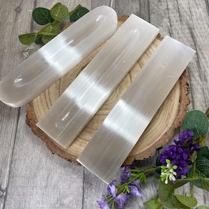 May include: Three selenite incense holders, two are rectangular and one is a curved oval shape. The holders are white and have a smooth, polished surface. They are arranged on a wooden surface.