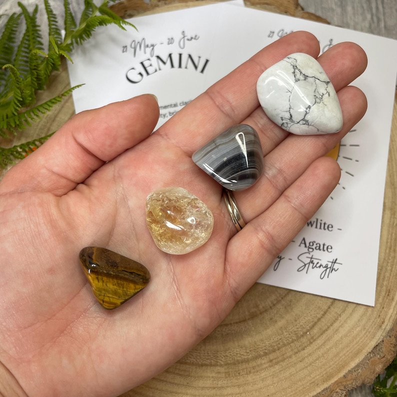 May include: A collection of polished gemstones in a hand, including a white howlite, a striped gray agate, a golden citrine, and a tiger's eye. A card with the word "Gemini" and the dates "May - June" is visible.