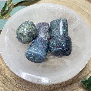May include: A selection of tumbled stones in shades of blue, green, and red, presented in a white, shallow bowl. The stones have a speckled appearance and various shapes. The bowl rests on a wooden surface, with greenery in the background.