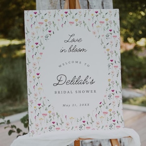 Può includere: Un cartello bianco su un cavalletto di legno, decorato con un bordo floreale. Il cartello recita "Love in bloom, Welcome to Delilah's Bridal Shower" con la data 21 maggio 20XX. Un tessuto bianco drappeggia la parte inferiore del cartello.