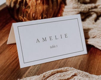 May include: White table card with black border and text "AMELIE table 1" in a minimalist font. The card is on a wooden surface with a beige fabric draped over it.
