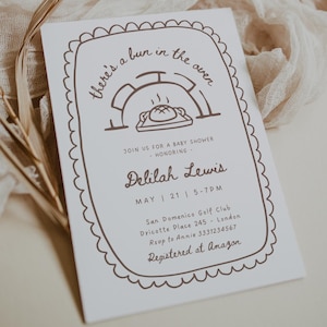 Può includere: Un invito per baby shower bianco con testo marrone e un bordo decorativo. Il testo recita "there's a bun in the oven" e invita gli ospiti a onorare Delilah Lewis. I dettagli includono data, ora e luogo.