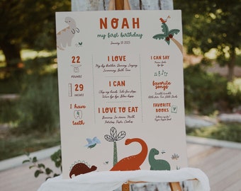 May include: A colourful first birthday sign for a boy named Noah. The sign features a dinosaur theme with illustrations of dinosaurs, a palm tree, and a bird. The sign includes information about Noah's age, weight, height, favourite foods, songs, and books.