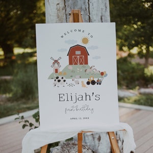 Puede incluir: Bienvenido al primer cumpleaños de Elijah, cartel con un granero rojo, animales de granja y un tractor. El cartel está en un caballete de madera y tiene un fondo blanco.
