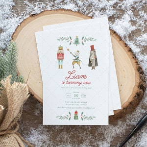 Può includere: Inviti bianchi con un'illustrazione ad acquerello di personaggi schiaccianoci e il testo "Liam compie un anno". Gli inviti sono su una fetta di legno, con un piccolo albero sempreverde e neve artificiale.