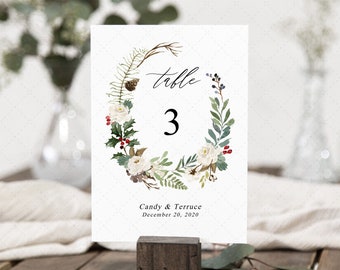 Pode incluir: Um cartão branco com número de mesa com um design de coroa de flores. A coroa inclui rosas brancas, folhas verdes e bagas vermelhas. A palavra "table" está escrita em itálico acima do número "3". Os nomes "Candy & Terruce" e a data "20 de dezembro de 2020" estão impressos na parte inferior.