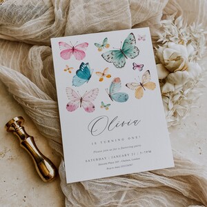 May include: A white invitation card with a butterfly theme. The card features a watercolor illustration of butterflies in various colors, including pink, blue, green, and yellow. The text on the card reads "Olivia is turning one!" and includes details about the party, such as the date, time, and location.