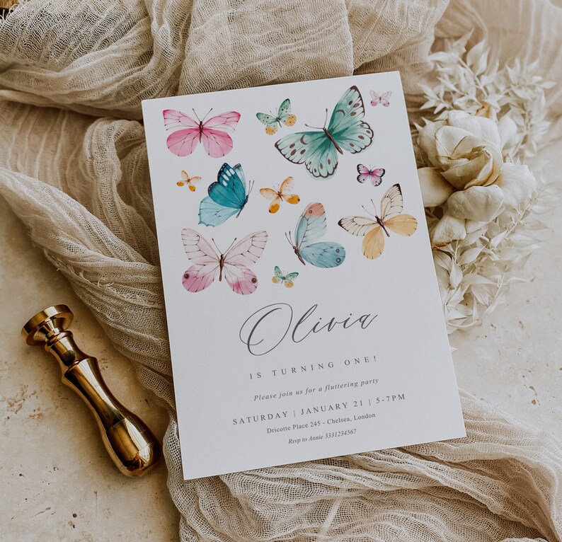 May include: A white invitation card with a butterfly theme. The card features a watercolor illustration of butterflies in various colors, including pink, blue, green, and yellow. The text on the card reads "Olivia is turning one!" and includes details about the party, such as the date, time, and location.