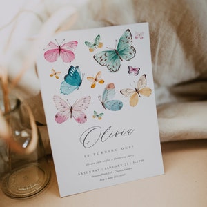 May include: A white invitation card with a butterfly theme. The card features a handwritten name "Olivia" and the text "Is turning one!" with details for a birthday party. The card is decorated with colorful watercolor butterflies.