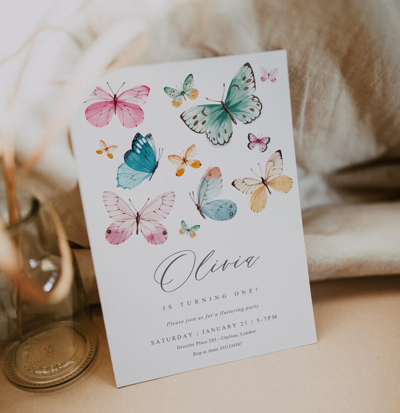 May include: A white invitation card with a butterfly theme. The card features a handwritten name "Olivia" and the text "Is turning one!" with details for a birthday party. The card is decorated with colorful watercolor butterflies.