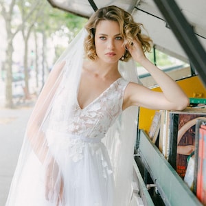 Peut inclure: Une mariée porte une robe de mariée blanche avec un décolleté en V et des détails de dentelle florale. Elle porte un voile transparent et a les cheveux blonds courts et bouclés. En arrière-plan, une librairie de rue.