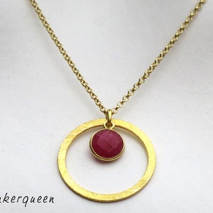 Könnte beinhalten: Eine goldene Halskette mit einem Kreisanhänger und einem roten Edelstein. Der Anhänger ist ein einfacher Kreis mit einer glatten, strukturierten Oberfläche. Der Edelstein ist ein runder, facettierter Stein, der in der Mitte des Kreises sitzt.