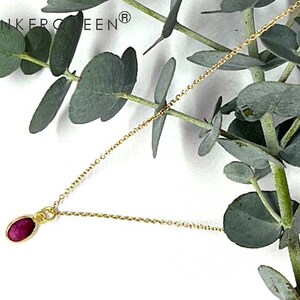 Könnte beinhalten: Eine zarte goldene Halskette mit einem kleinen, oval geformten Rubinanhänger. Der Anhänger hat eine leuchtend rote Farbe und ist in eine goldene Lünette gefasst. Die Halskette wird auf einem Zweig Eukalyptus mit grünen Blättern präsentiert.