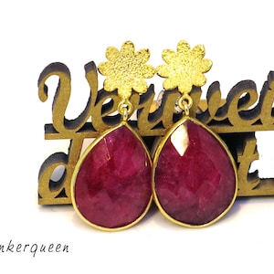 Könnte beinhalten: Ein Paar Ohrringe mit einem tropfenförmigen rubinroten Edelstein und einem goldenen Blumenanhänger.