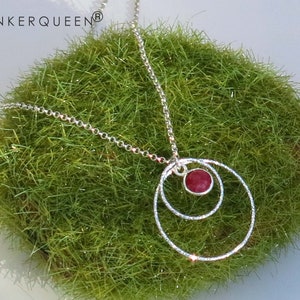 Könnte beinhalten: Eine silberne Halskette mit einem roten Edelstein-Anhänger. Der Anhänger ist von zwei silbernen Kreisen umgeben.
