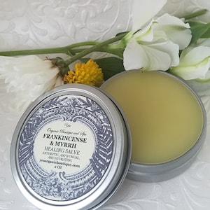 May include: A silver tin with a white label that reads "Yos Organic Boutique and Spa Frankincense & Myrrh Healing Salve Antiseptic, Antifungal, and Hydrating yesorganicboutique.com 2 OZ". The tin is open and contains a yellow salve.