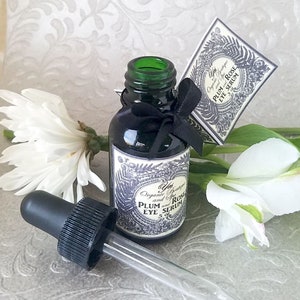 Pode incluir: Uma garrafa de vidro verde escuro de s&eacute;rum para os olhos com um conta-gotas preto e uma etiqueta. A etiqueta diz "Plum and Rose Eye Serum". Uma fita preta est&aacute; amarrada ao redor da garrafa. Flores brancas no fundo.