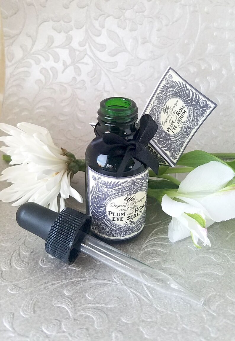Pode incluir: Uma garrafa de vidro verde escuro de s&eacute;rum para os olhos com um conta-gotas preto e uma etiqueta. A etiqueta diz "Plum and Rose Eye Serum". Uma fita preta est&aacute; amarrada ao redor da garrafa. Flores brancas no fundo.