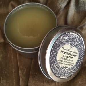 May include: A silver tin with a label that reads "Organic Boutique and Spa Frankincense & Myrrh Healing Salve Antiseptic and healing to dry, cracked, and scarred skin 3 oz". The tin is open and filled with a light yellow salve.