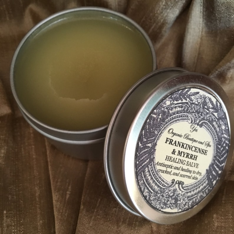 May include: A silver tin with a label that reads "Organic Boutique and Spa Frankincense & Myrrh Healing Salve Antiseptic and healing to dry, cracked, and scarred skin 3 oz". The tin is open and filled with a light yellow salve.