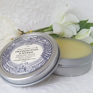 May include: A silver tin with a label that reads "Frankincense & Myrrh Healing Salve Antiseptic, Antifungal, and Hydrating 2 oz ysorganicboutique.com". The tin is open and the salve is a pale yellow color.