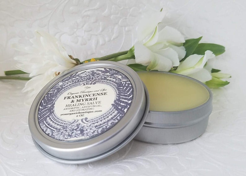 May include: A silver tin with a label that reads "Frankincense & Myrrh Healing Salve Antiseptic, Antifungal, and Hydrating 2 oz ysorganicboutique.com". The tin is open and the salve is a pale yellow color.