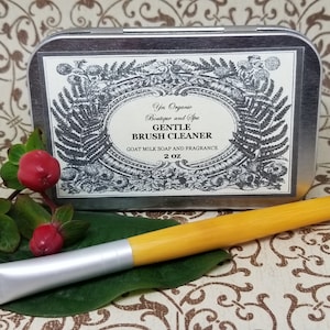 Op de afbeelding: Een zilveren blik met een zwart-wit etiket waarop staat "Yes Organic Boutique and Spa Gentle Brush Cleaner Goat Milk Soap and Fragrance 2 oz". Het blik staat op een bruin-wit gepatroneerd oppervlak naast een gele en zilveren make-upborstel.