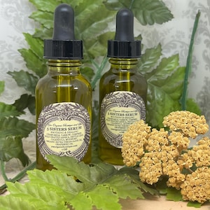 May include: Two amber glass bottles with black dropper tops filled with a yellow liquid. The bottles are labeled "5 Sisters Serum" and list ingredients including chamomile, calendula, and wildcrafted arnica. The bottles are sitting on a wooden surface with green foliage and dried flowers.