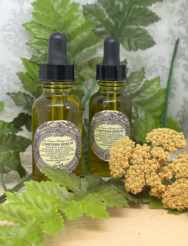 May include: Two amber glass bottles with black dropper tops filled with a yellow liquid. The bottles are labeled "5 Sisters Serum" and list ingredients including chamomile, calendula, and wildcrafted arnica. The bottles are sitting on a wooden surface with green foliage and dried flowers.