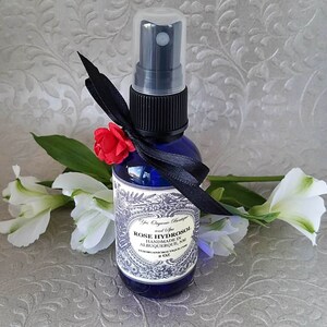 Puede incluir: Una botella de vidrio azul de 2 oz con un atomizador negro y una cinta negra atada alrededor del cuello. La botella contiene hidrosol de rosa y tiene una etiqueta que dice "Rose Hydrosol Handmade in Albuquerque, NM  YosOrganicBoutique.com".