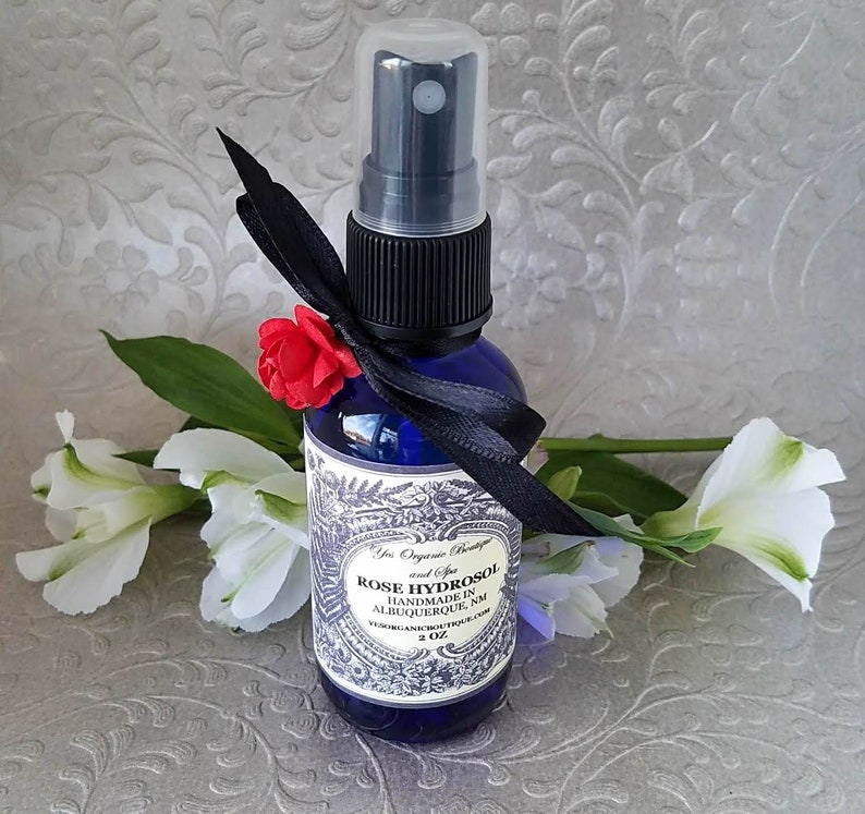 Puede incluir: Una botella de vidrio azul de 2 oz con un atomizador negro y una cinta negra atada alrededor del cuello. La botella contiene hidrosol de rosa y tiene una etiqueta que dice "Rose Hydrosol Handmade in Albuquerque, NM  YosOrganicBoutique.com".