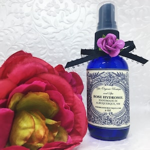 Op de afbeelding: Een blauwe glazen fles met een zwart-wit etiket waarop staat "Yes Organic Boutique and Spa Rose Hydrosol Handmade in Albuquerque, NM yesorganicboutique.com 2 oz". De fles heeft een zwarte spuitdop en een roze roos bovenop de fles.