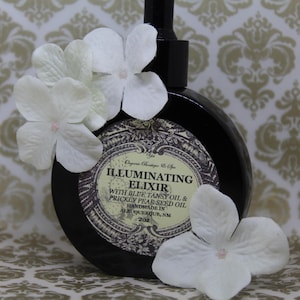 Puede incluir: Una botella de vidrio oscuro con una etiqueta blanca que dice "Illuminating Elixir with Blue Tansy Oil & Prickly Pear Seed Oil Handmade in Albuquerque, NM 208". La botella está rodeada de flores blancas.