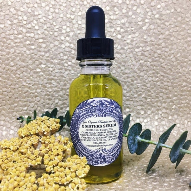 May include: A clear glass bottle with a black dropper top filled with a yellow liquid. The bottle has a white label with blue and gold accents. The label reads "5 Sisters Serum" and lists the ingredients: chamomile, yarrow, comfrey, wild crafted arnica, nettle leaf, calendula, mugwort, organic extra virgin olive oil, grapefruit oil, aroma. The bottle is sitting on a gold background with yellow flowers and green leaves.
