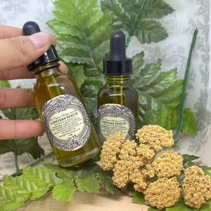 May include: Two amber glass bottles with dropper tops filled with a golden liquid. The bottles are labeled "5 Sisters Serum" and "5 Sisters Serum" with a list of ingredients. The bottles are sitting on a surface with green leaves and dried flowers.
