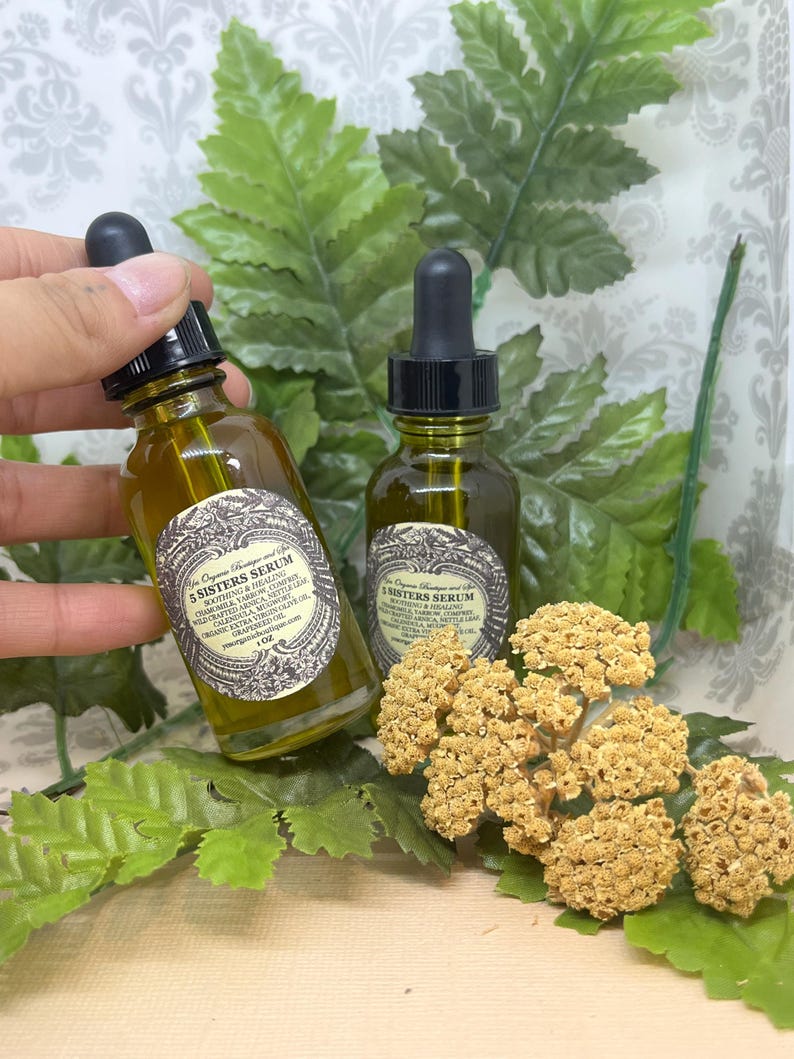 May include: Two amber glass bottles with dropper tops filled with a golden liquid. The bottles are labeled "5 Sisters Serum" and "5 Sisters Serum" with a list of ingredients. The bottles are sitting on a surface with green leaves and dried flowers.