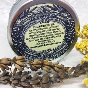 May include: A silver tin with a black and white label that reads "INGREDIENTS: GRAPESEED OIL INFUSED WITH MYRRH GUM PIECES AND FRANKINCENSE TEARS, LOCAL BEESWAX, FRANKINCENSE AND MYRRH ESSENTIAL OILS HANDMADE IN ALBUQUERQUE, NM yesorganicboutique.com"