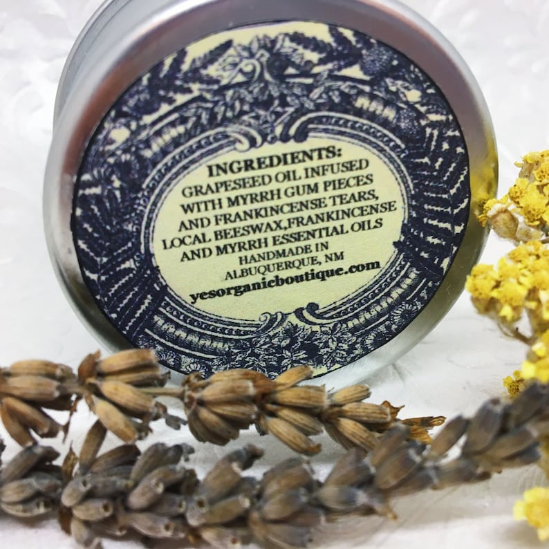 May include: A silver tin with a black and white label that reads "INGREDIENTS: GRAPESEED OIL INFUSED WITH MYRRH GUM PIECES AND FRANKINCENSE TEARS, LOCAL BEESWAX, FRANKINCENSE AND MYRRH ESSENTIAL OILS HANDMADE IN ALBUQUERQUE, NM yesorganicboutique.com"