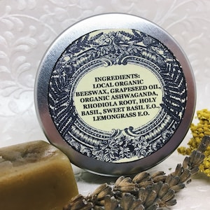 May include: A silver tin with a blue and white label listing the ingredients: Local Organic Beeswax, Grapeseed Oil, Organic Ashwaganda, Rhodiola Root, Holy Basil, Sweet Basil E.O., Lemongrass E.O.