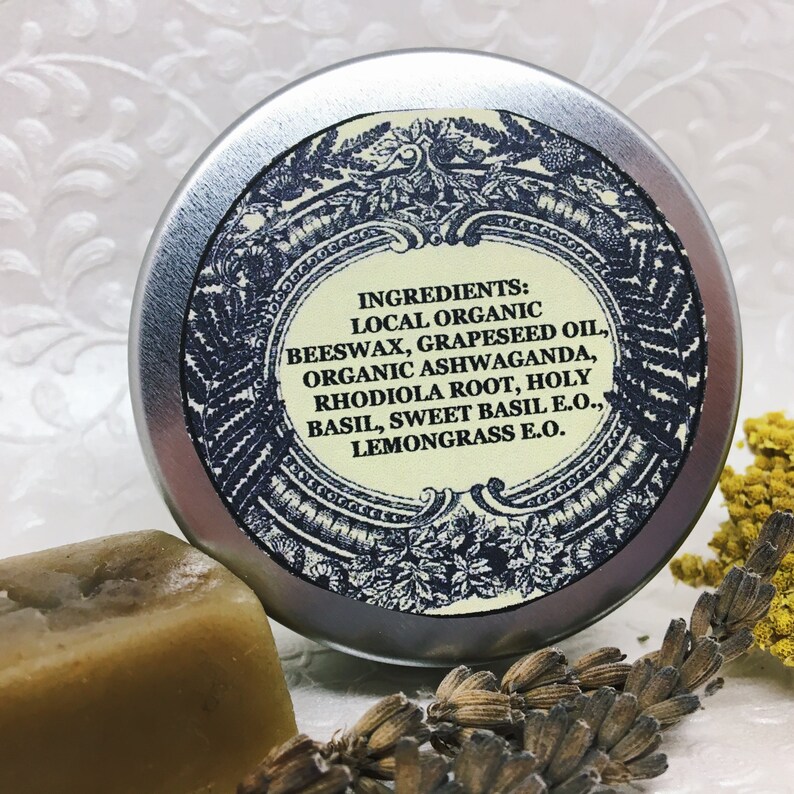 May include: A silver tin with a blue and white label listing the ingredients: Local Organic Beeswax, Grapeseed Oil, Organic Ashwaganda, Rhodiola Root, Holy Basil, Sweet Basil E.O., Lemongrass E.O.