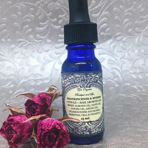 May include: A blue glass bottle with a dropper top containing Frankincense & Myrrh Cuticle + Nail Growth Oil. The label is white with black text and a decorative border. The bottle is sitting on a surface with dried pink roses.