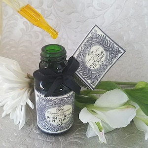 Pode incluir: Uma garrafa de vidro verde escuro de s&eacute;rum para os olhos com um la&ccedil;o preto, um conta-gotas com l&iacute;quido amarelo e uma caixa pequena. O r&oacute;tulo diz "Plum and Rose Eye Serum". Flores brancas ao fundo.