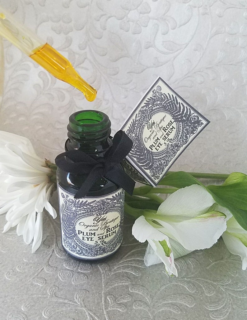 Pode incluir: Uma garrafa de vidro verde escuro de s&eacute;rum para os olhos com um la&ccedil;o preto, um conta-gotas com l&iacute;quido amarelo e uma caixa pequena. O r&oacute;tulo diz "Plum and Rose Eye Serum". Flores brancas ao fundo.