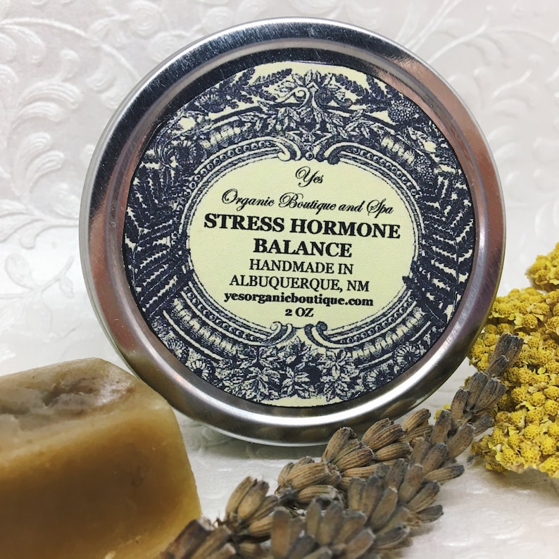 May include: A silver tin with a black and white label that reads "Yes Organic Boutique and Spa Stress Hormone Balance Handmade in Albuquerque, NM yesorganicboutique.com 2 OZ".