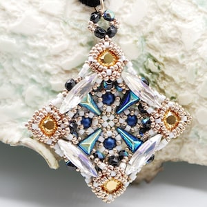 May include: A square-shaped pendant with a geometric design featuring white, blue, gold, and iridescent beads. The pendant is attached to a black cord.