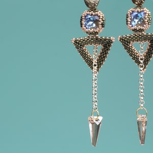 May include: A pair of dangle earrings with a geometric triangle design. The earrings are made with black and gold beads and feature a blue crystal in the center of each triangle. The earrings have a silver chain with a clear crystal at the end.