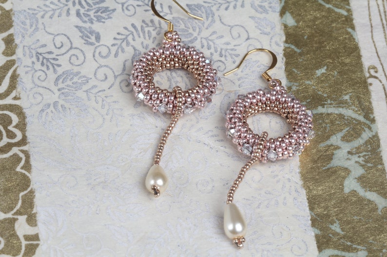 Op de afbeelding: Een paar gouden oorbellen met roze en heldere kralen, elk met een parel hanger.