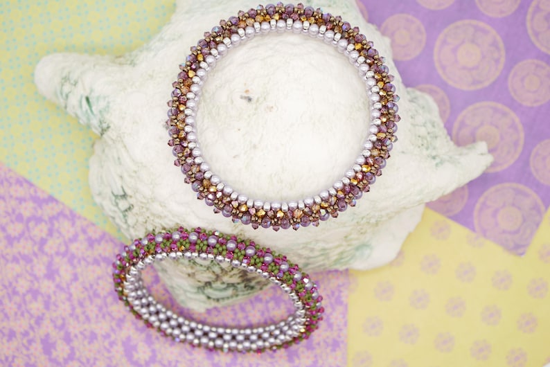 May include: Two beaded bracelets, one with purple and gold beads and the other with green and purple beads, are displayed on a white seashell.
