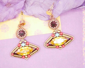 May include: A pair of handmade earrings featuring a geometric design. The earrings have a gold-tone metal frame, with a large yellow gemstone in the center. The design incorporates red, purple, and turquoise beads. The earrings are displayed on a purple background.
